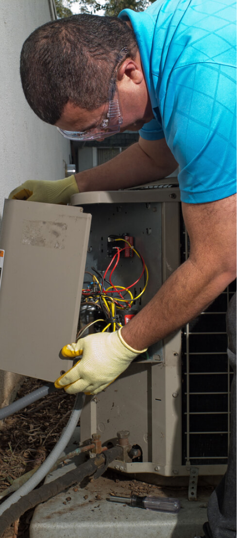 A technician in a blue shirt and safety glasses opens the access panel of an outdoor air conditioning unit to perform repairs.