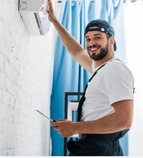 A smiling technician in a uniform and cap installs a wall-mounted air conditioning unit while holding a screwdriver.