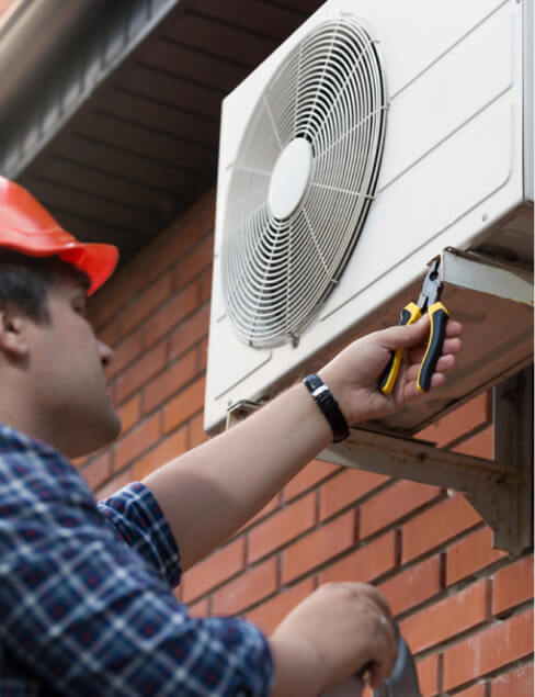 A technician in a hard hat uses pliers to repair an outdoor air conditioning unit mounted on a brick wall.