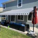 A striped retractable awning shades a patio with outdoor furniture, a grill, and a closed red umbrella beside a gray house on a sunny day.