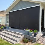 A porch with a dark retractable screen covering its open side, surrounded by potted plants and a landscaped yard.