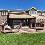 Two-story brick house with solar panels, beige awnings, and a wooden deck facing a green lawn under a clear blue sky.