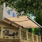 A beige and white striped retractable awning extends over a wooden deck attached to a house, surrounded by trees and greenery.
