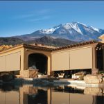 A house with tan retractable patio screens, a swimming pool in the foreground, and a snow-capped mountain in the background under a clear blue sky.