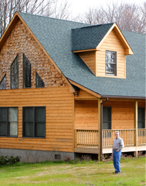 A man standing in the yard of a wooden house with a green shingled roof.