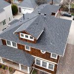 Aerial view of a two-story house with a gray shingle roof, brown siding, a front porch, driveway, and nearby parked cars in a residential neighborhood.