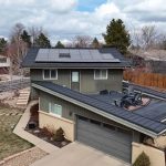 Two-story house with solar panels on the roof, surrounded by a brown wooden fence and sparse trees, photographed on a partly cloudy day.