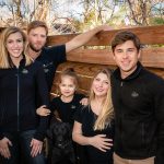 Five people and a black dog pose together outdoors by a wooden fence, all wearing black shirts or jackets with a matching green logo. Trees are visible in the background.
