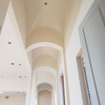 Arched hallway with high vaulted ceilings, cream-colored walls, and recessed lighting, featuring a tall window on the right.