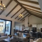 Open-concept living room and kitchen with vaulted ceiling, exposed wooden beams, large windows, modern chandeliers, and neutral-colored furniture.