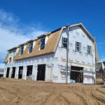 A partially constructed two-story building wrapped in Tyvek sheeting stands on a dirt lot under a blue sky. Construction materials are visible nearby.