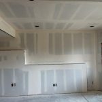 Unpainted drywall with visible seams and electrical outlets in a room under construction, featuring a window and unfinished floor.