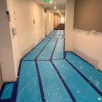 A hallway under renovation with blue protective floor covering and tape, unfinished walls, and a lit exit sign at the end.