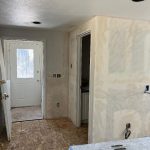 Room under renovation with unfinished drywall, exposed subfloor, and uninstalled electrical outlets; a door and open doorway are visible.
