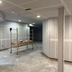 Unfinished basement room with drywall installed, a scaffolding cart, construction tools, and concrete floors visible under bright ceiling lights.
