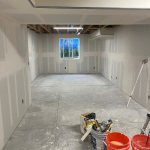 Unfinished room with drywall installed, tools and red buckets on the floor, and a window at the far end showing a view outside.