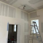 A room under construction with unfinished drywall, a stepladder, open doorway, and supplies visible in the adjacent room. Two ceiling light fixture openings are exposed.