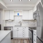 Modern kitchen with white cabinets, stainless steel appliances, mosaic tile backsplash, and a blue vase on a shelf above the sink. Recessed lighting and wood-look flooring are visible.
