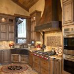 Rustic kitchen with wood cabinets, tile floor, a large range hood, double ovens, farmhouse sink, and a window over the sink. A round rug is on the floor in front of the stove.