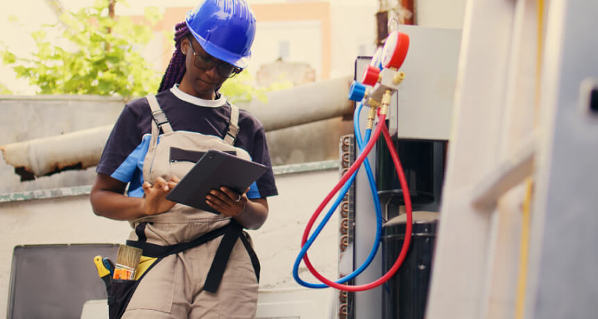 Person wearing overalls and a hard hat writes on a clipboard near HVAC equipment outdoors, with tools and colored hoses visible.