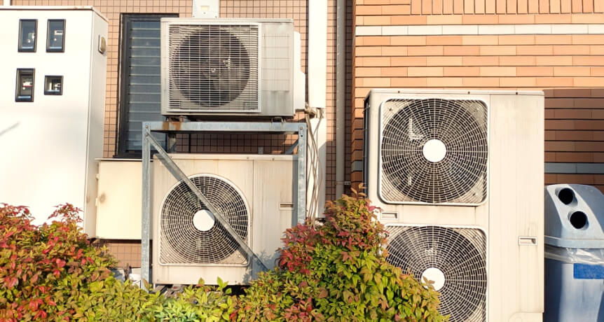 Four outdoor air conditioning units are installed against a brick wall. Shrubs are in the foreground, and a blue-gray trash bin is on the right.