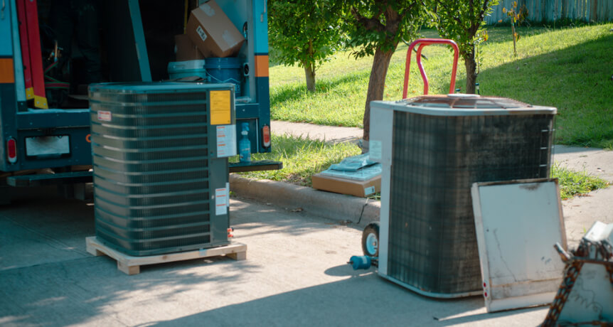 Two air conditioning units are on the ground near a service truck, with equipment and tools scattered around, suggesting installation or maintenance work.